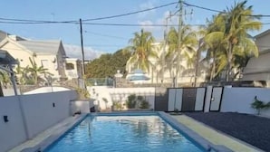 A rectangular swimming pool with a stone border, surrounded by a low wall and palm trees. - Splendid 4-bedrooms villa private pool near beach (Flic en Flac)