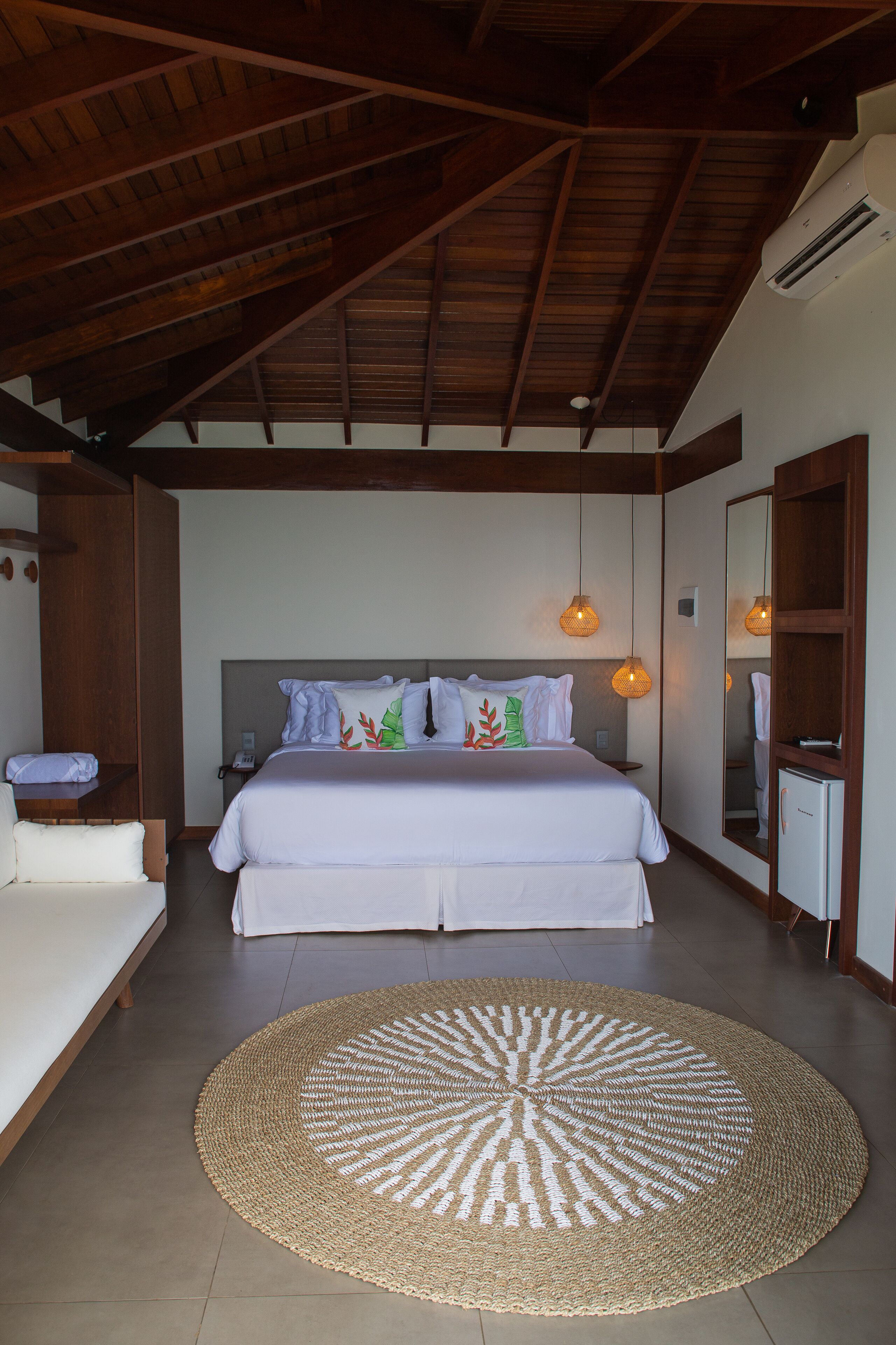 A bedroom with a large bed, a round woven rug, wooden beams on the ceiling, and a mirror on the wall.