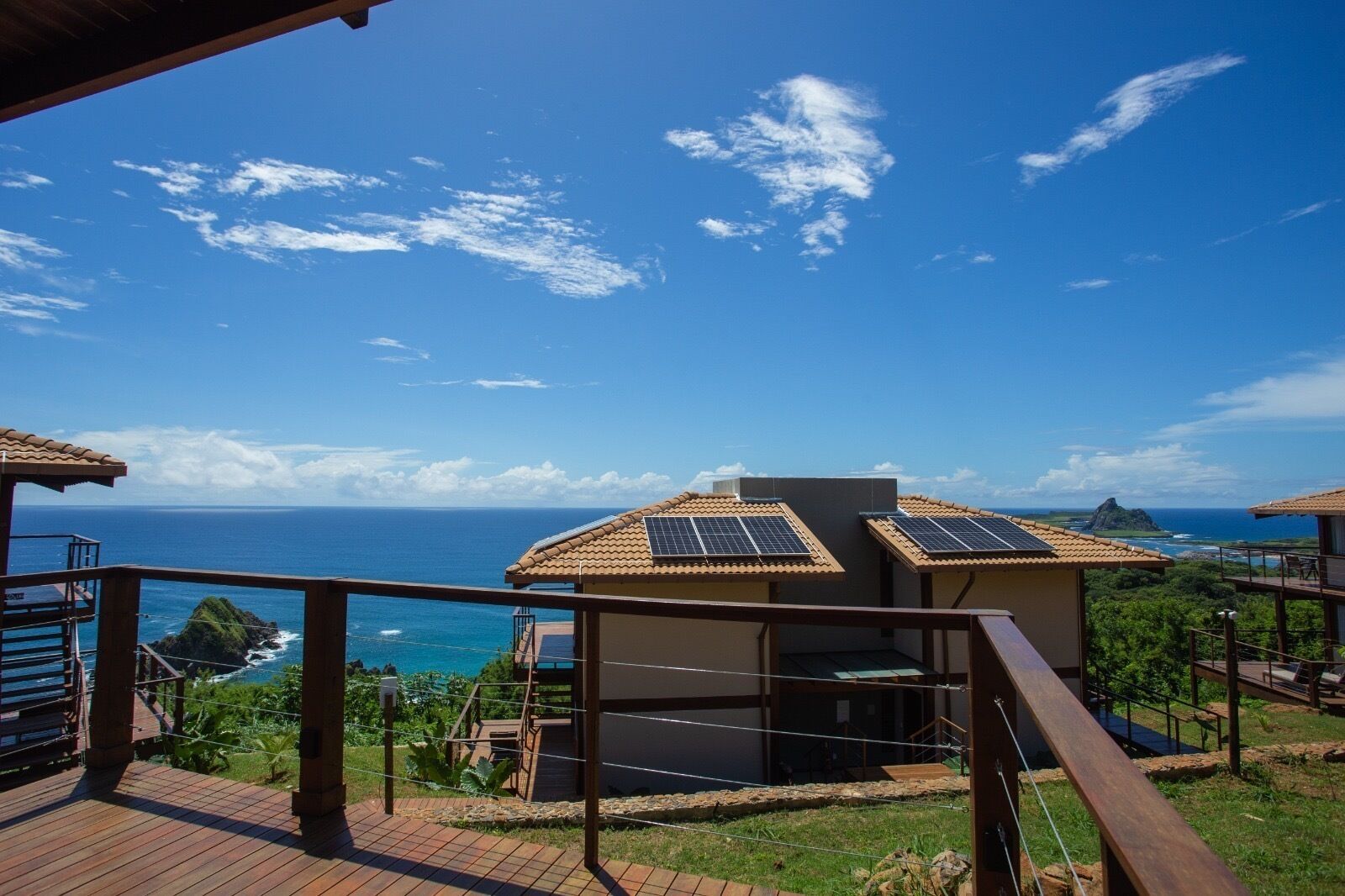 A coastal property with a wooden deck, solar panels on the roof, and a view of the ocean and a mountain.