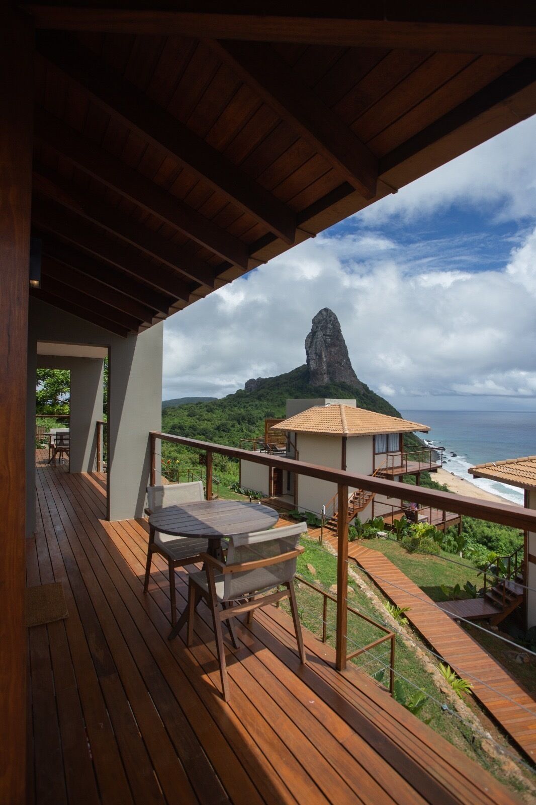 A wooden deck with a table and chairs, overlooking a beach and a mountain.