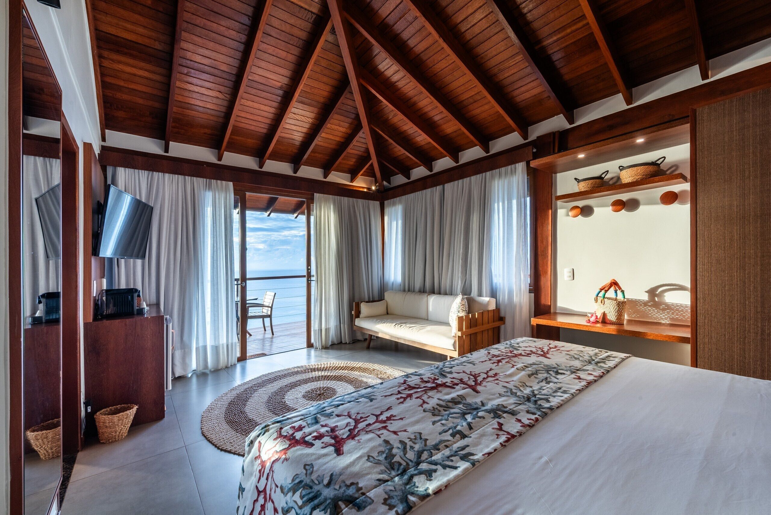 Un dormitorio con cama, sofá, televisor, techo de madera y vista al mar.