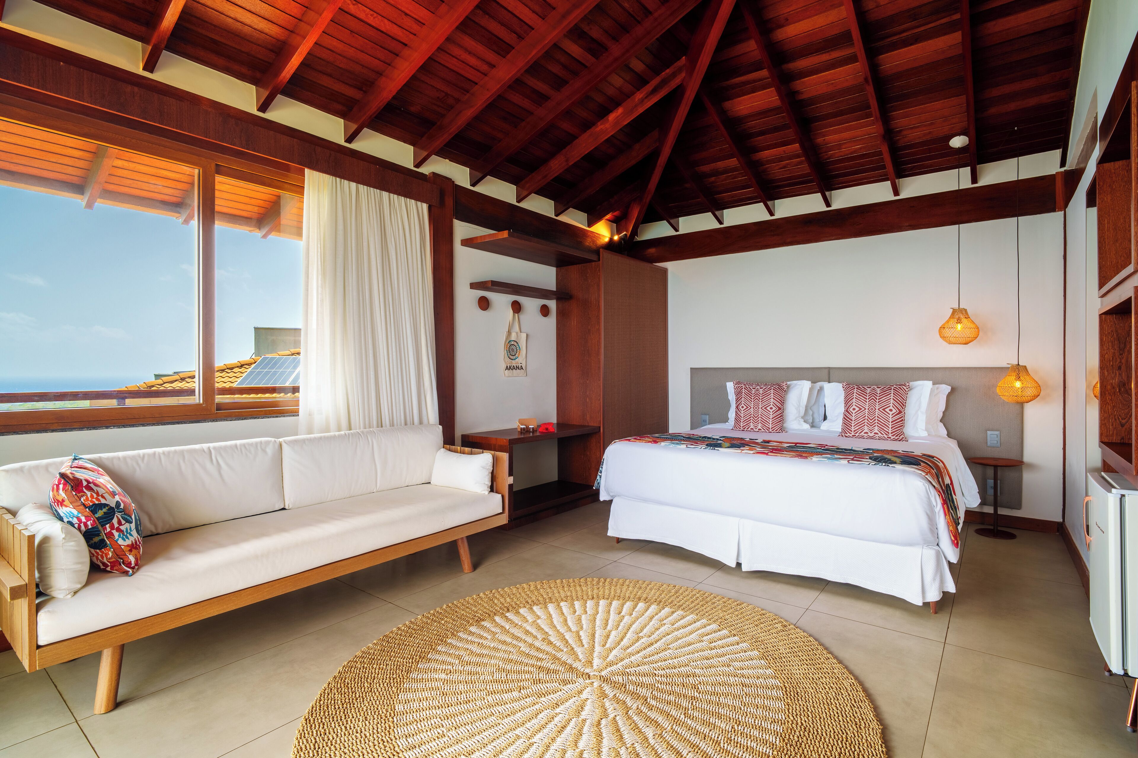 A bedroom with a bed, a sofa, a round woven rug, and a view of the sea.