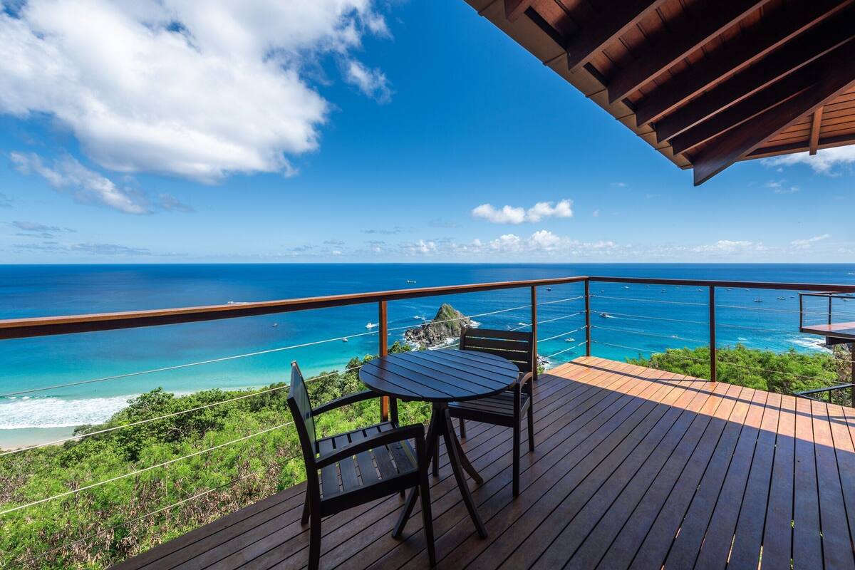 A wooden deck with two chairs and a table overlooking a coastal view with boats and a rocky island.