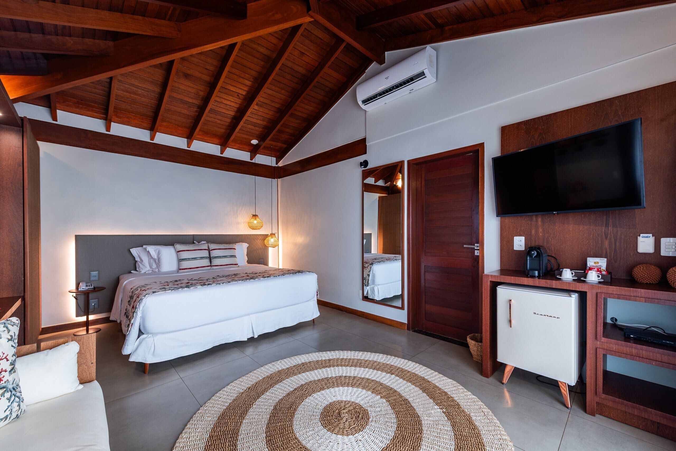 A bedroom with a bed, bedside table, lamp, air conditioning unit, television, and a small refrigerator.