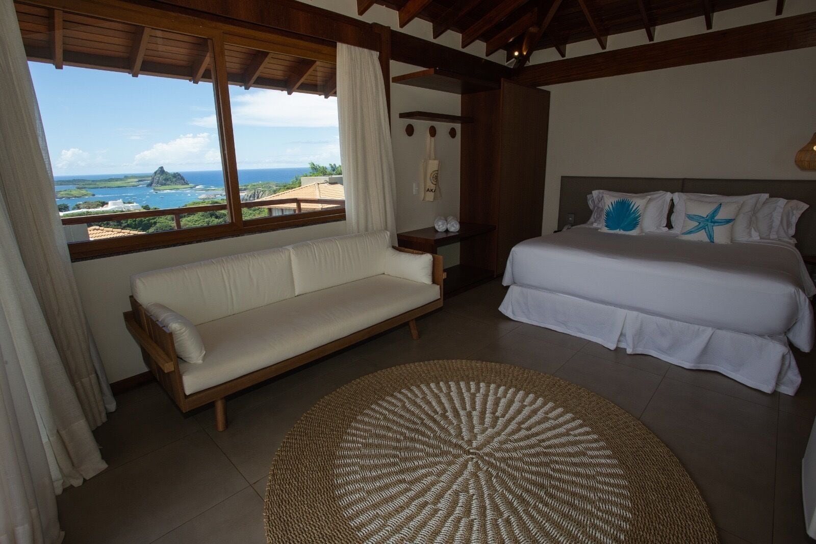 A bedroom with a bed, a sofa, a round woven rug, and a view of the ocean.