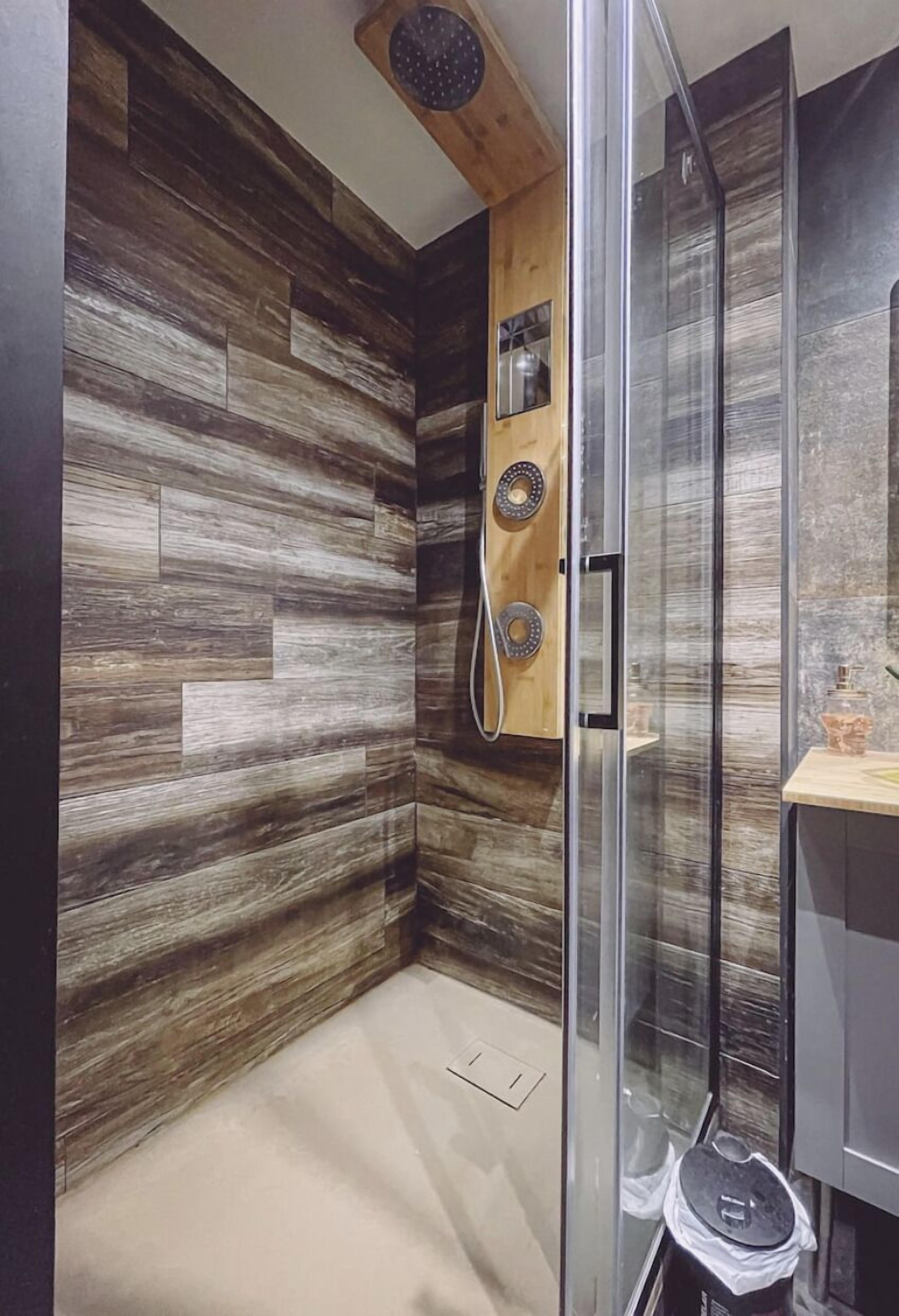 A modern bathroom with a wooden wall panel design, a glass shower enclosure, and a small wooden shelf with a mirror and a bottle.
