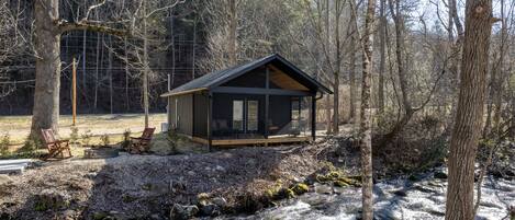 Una cabaña con terraza y dos sillas, rodeada de un bosque y un arroyo.