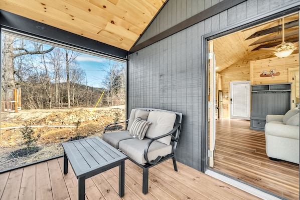 A wooden deck with a sofa and table, a large window, and a view of a garden.
