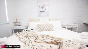 Ein Schlafzimmer mit weißer Backsteinwand, einem Bett mit gemustertem Überwurf, zwei Nachttischen mit Büchern und einer kleinen Lampe sowie einem gerahmten Bild an der Wand.