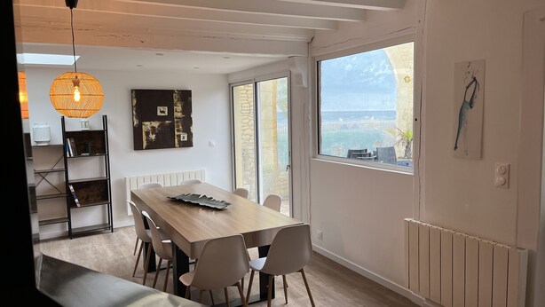 Een moderne eethoek met een houten tafel en stoelen, een hanglamp en uitzicht op de zee door grote ramen.
