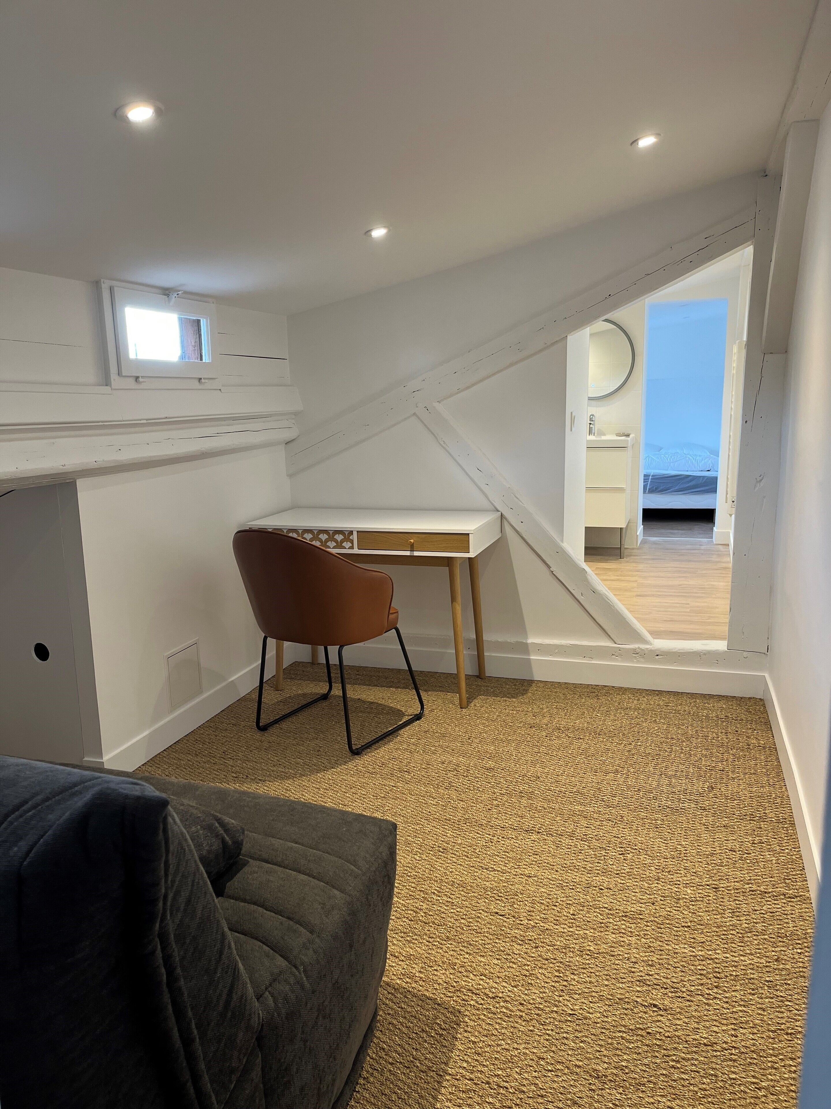 A compact, well-lit attic bedroom with a sloped ceiling, a small desk, a chair, and a view into an adjacent bathroom.