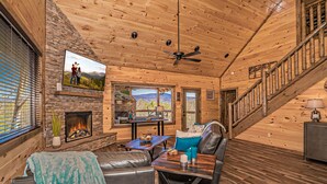 A cozy cabin interior with a stone fireplace, leather seating, a dining area, and a staircase. - To Infinity And Beyond: Indoor-Pool, VIEWS, Theater, Game Room, Hot Tub, WiFi, close to Attractions! (Sevierville)
