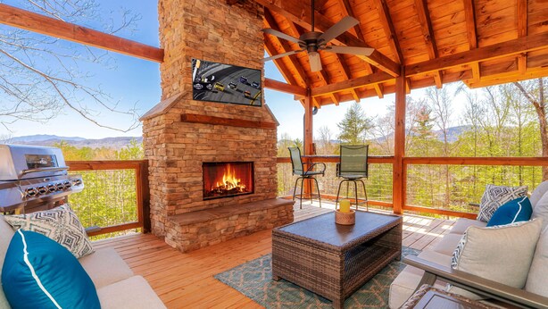 A covered outdoor area with a stone fireplace, wooden furniture, and a view of the surrounding landscape. - To Infinity And Beyond: Indoor-Pool, VIEWS, Theater, Game Room, Hot Tub, WiFi, close to Attractions! (Sevierville)