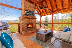 A covered outdoor area with a stone fireplace, wooden furniture, and a view of the surrounding landscape. - To Infinity And Beyond: Indoor-Pool, VIEWS, Theater, Game Room, Hot Tub, WiFi, close to Attractions! (Sevierville)