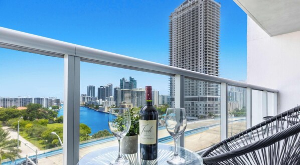 A balcony with a city view, a bottle of wine, and two glasses on a table. - Pool | Apartment w/ Balcony - Gym - Near Beach (Hallandale Beach)