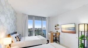 A hotel room with two beds, a desk, a chair, a balcony with a view of the city, and a painting on the wall. - Pool | Apartment w/ Balcony - Gym - Near Beach (Hallandale Beach)