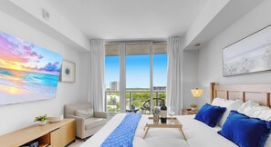 A hotel room with a large window, a bed with blue pillows, a bedside table with a wine glass, and a painting of a sunset. - Pool | Apartment w/ Balcony - Gym - Near Beach (Hallandale Beach)