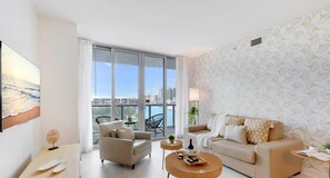 A modern living room with a large window, a sofa, a coffee table, and a dining table. - Pool | Apartment w/ Balcony - Gym - Near Beach (Hallandale Beach)