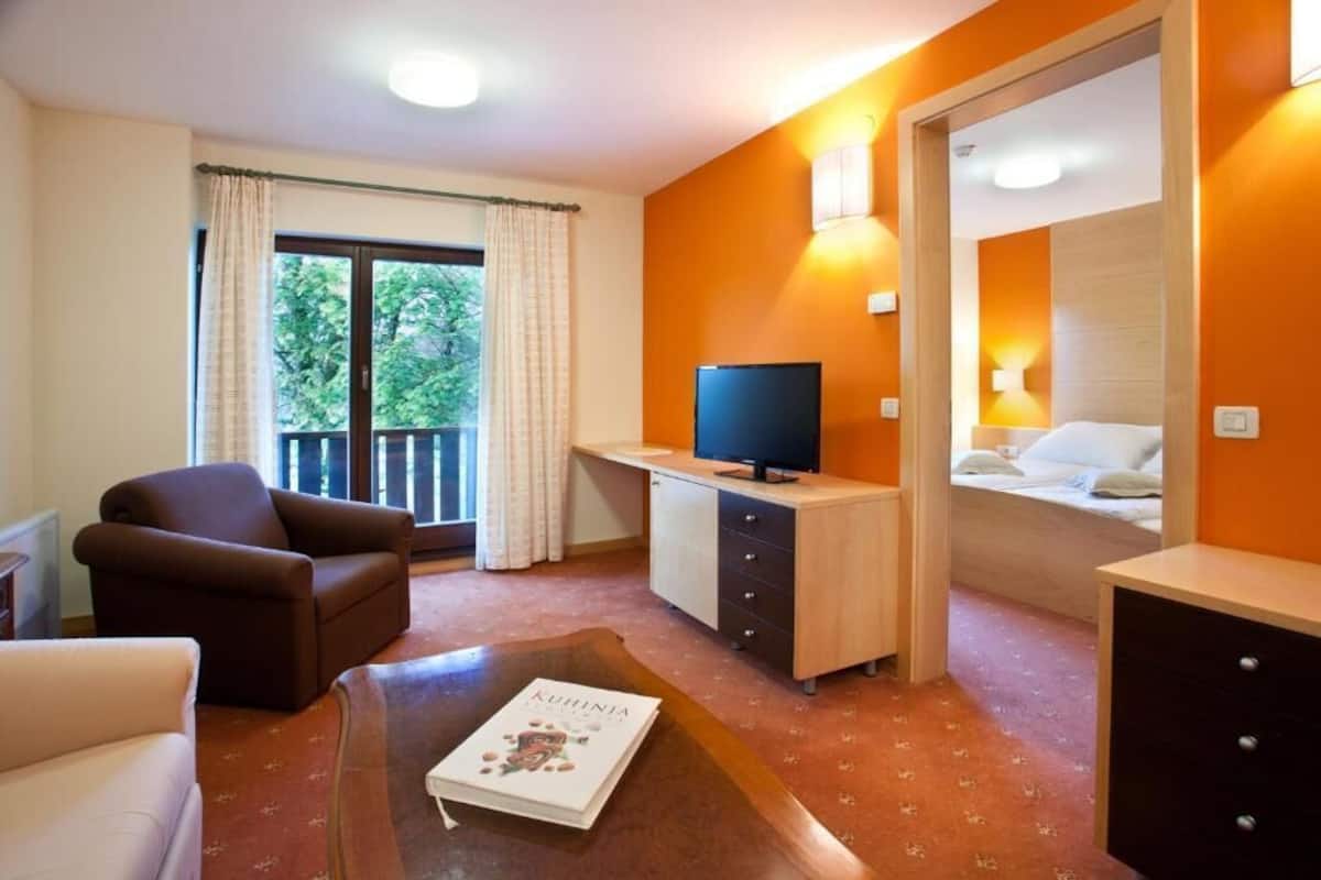 Ein Hotelzimmer mit einer Couch, einem Fernseher auf einer Holzkommode, einem Buch auf einem Couchtisch und einem Balkon mit Vorhängen.