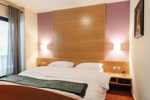Una habitación de hotel con cabecera de madera, una cama con ropa blanca, dos lámparas de noche y una mesita de noche con una planta.