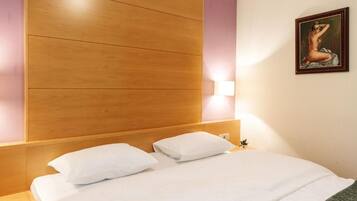 Habitación de hotel con cabecera de madera, ropa de cama blanca, luz montada en la pared y un cuadro enmarcado.