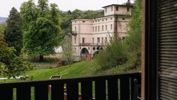 A large, historic building with multiple stories and arched windows, surrounded by lush greenery and trees.
