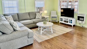 Una sala de estar con un sofá seccional gris, un sillón blanco, una mesita pequeña blanca y un televisor sobre un mueble blanco.