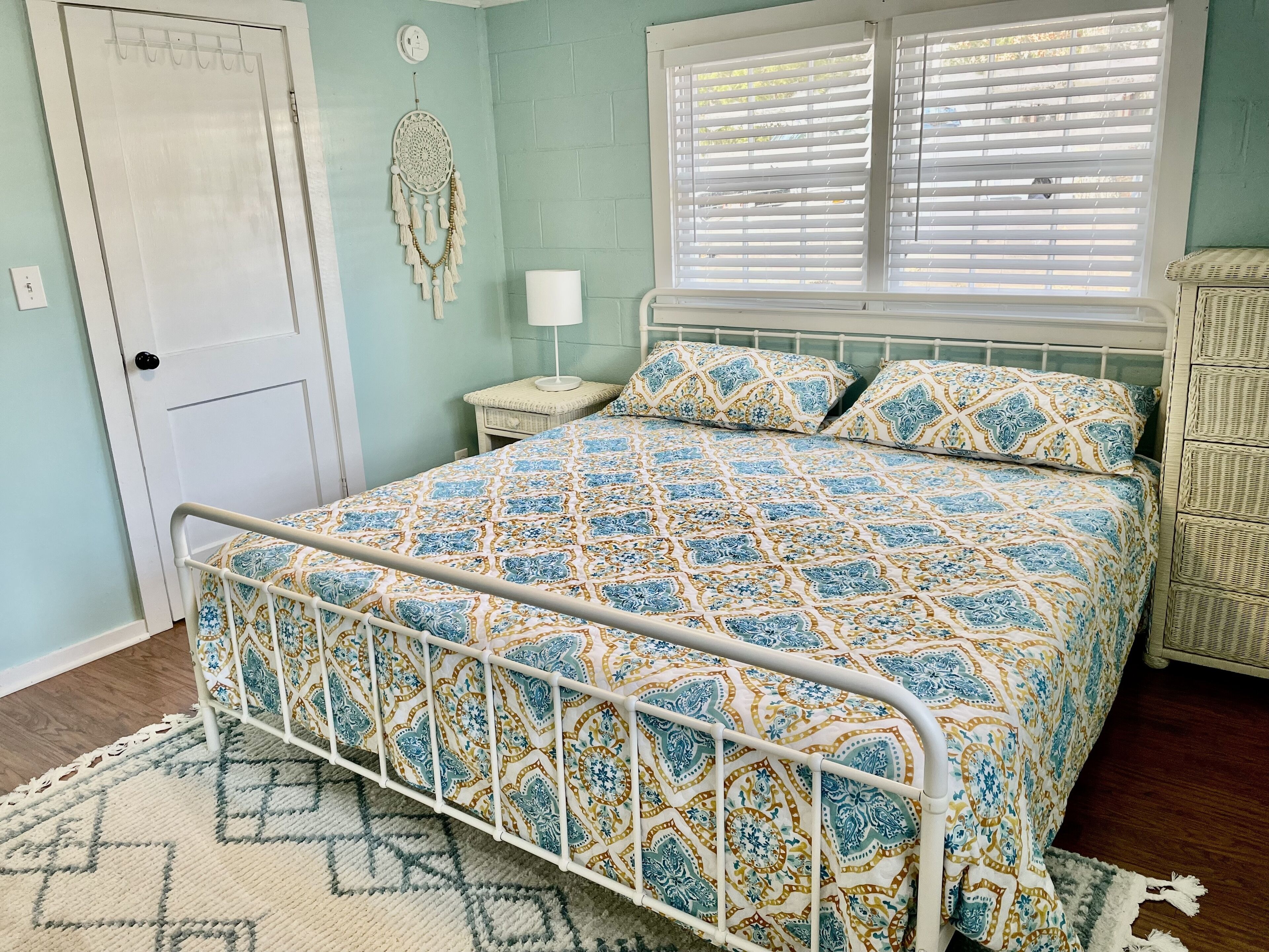 Une chambre avec une couvre-lit à motifs, un cadre de lit en métal blanc, une table de chevet avec une lampe et un attrape-rêves fixé au mur.
