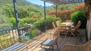 Una terraza de madera con mobiliario de mimbre y una mesa, con vistas a un paisaje verde exuberante y una piscina.