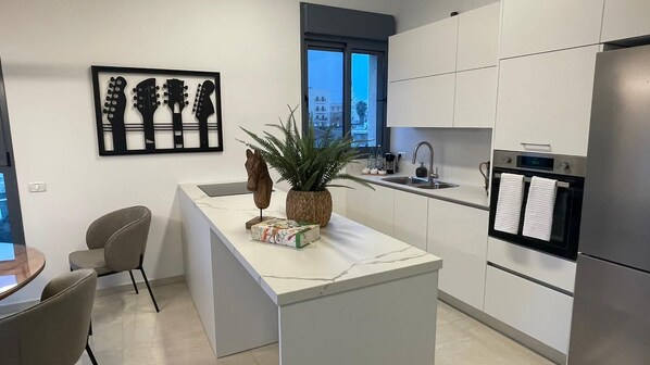 A modern kitchen with white cabinets, a built-in oven, and a sink. There is a potted plant on the island and a framed artwork on the wall.