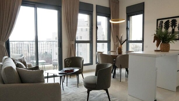 Una sala de estar moderna con sofá, sillones, una mesita y un comedor. Amplias ventanas ofrecen luz natural y vistas de la ciudad.