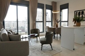 A modern living room with a sofa, chairs, a small table, and a dining area. Large windows provide natural light and a view of the city.