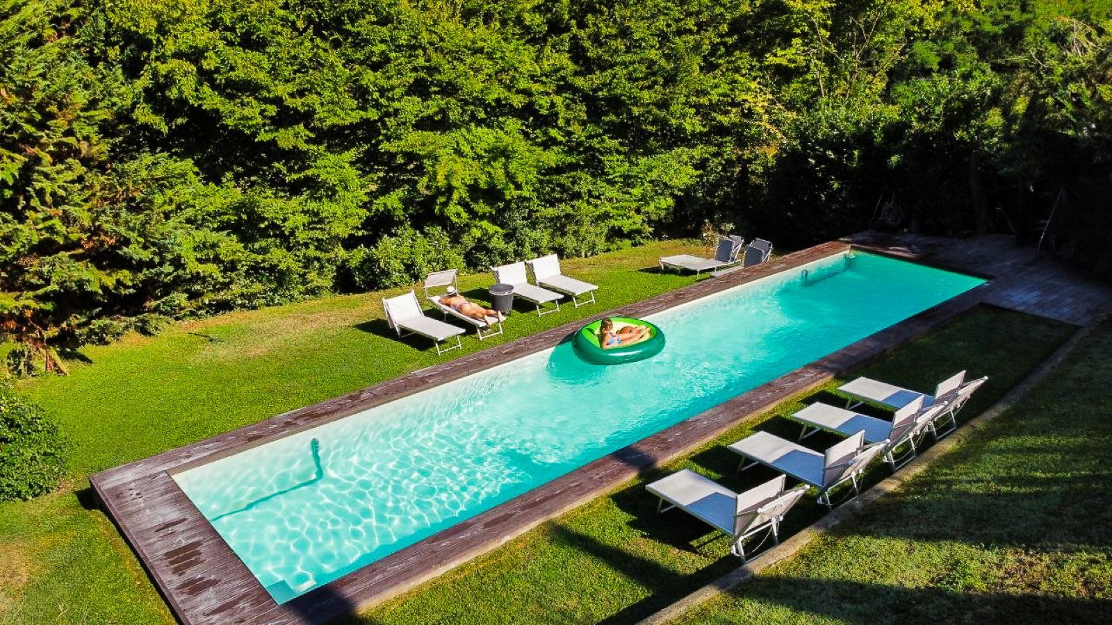 A rectangular swimming pool with a wooden deck surrounding it, a lounge area with chairs, and a grassy area with trees in the background.
