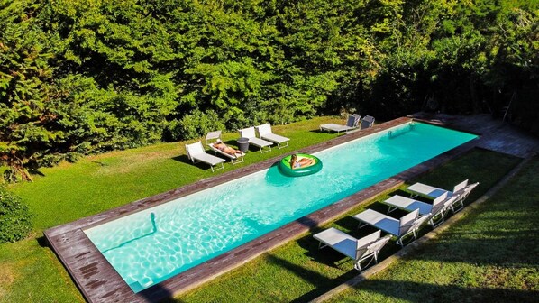A rectangular swimming pool with a wooden deck surrounding it, a lounge area with chairs, and a grassy area with trees in the background.