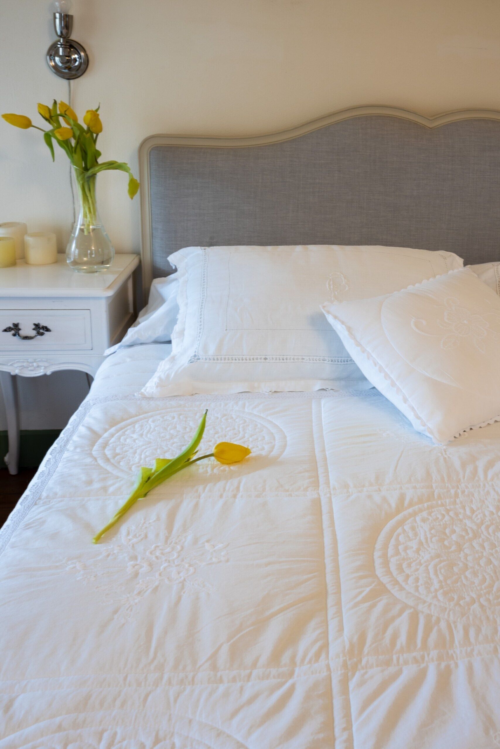 Una cama bien hecha con sábanas blancas y una única tulipán amarilla sobre el edredón.
