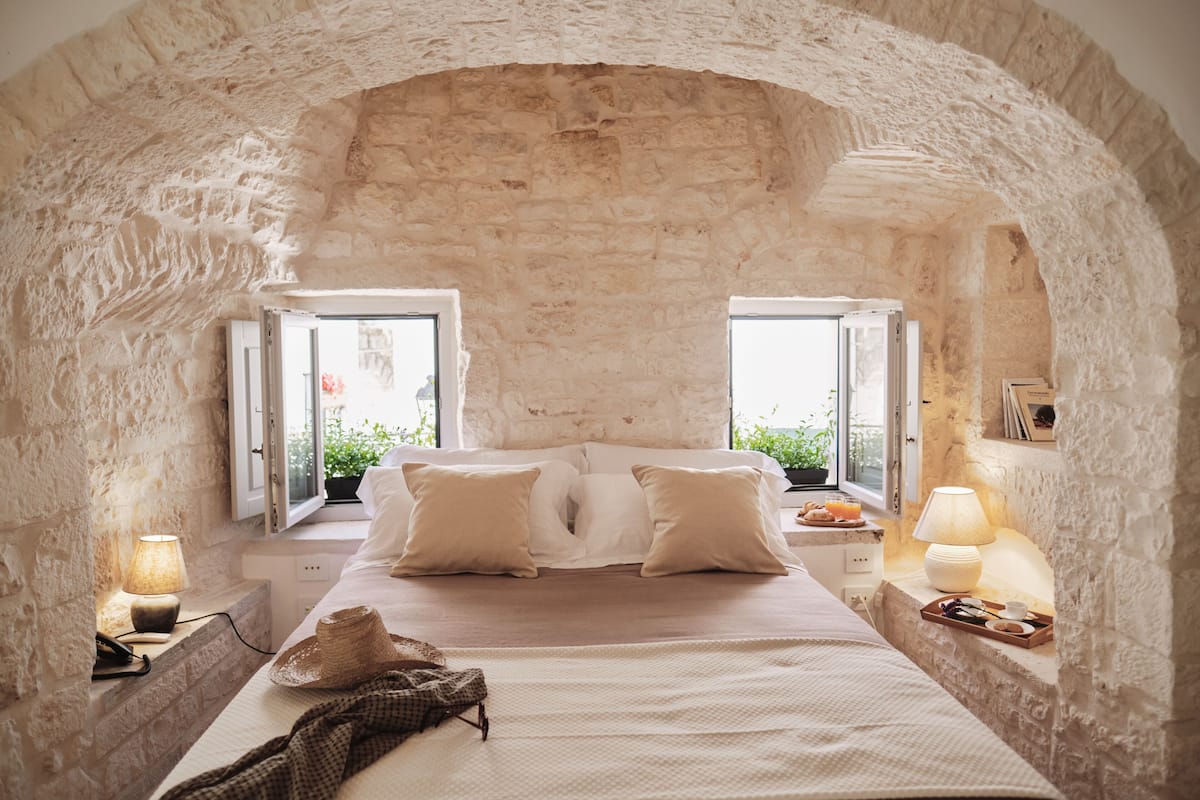 A bedroom with a stone arch, a bed with white linens, two bedside tables with lamps, and a window with a view of greenery.