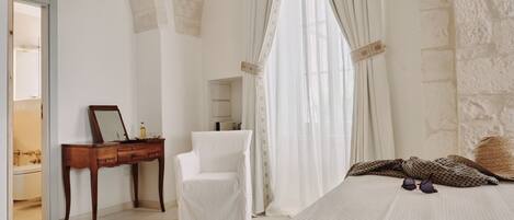 Un dormitorio con cama, una silla, tocador con espejo y ventana con cortinas.