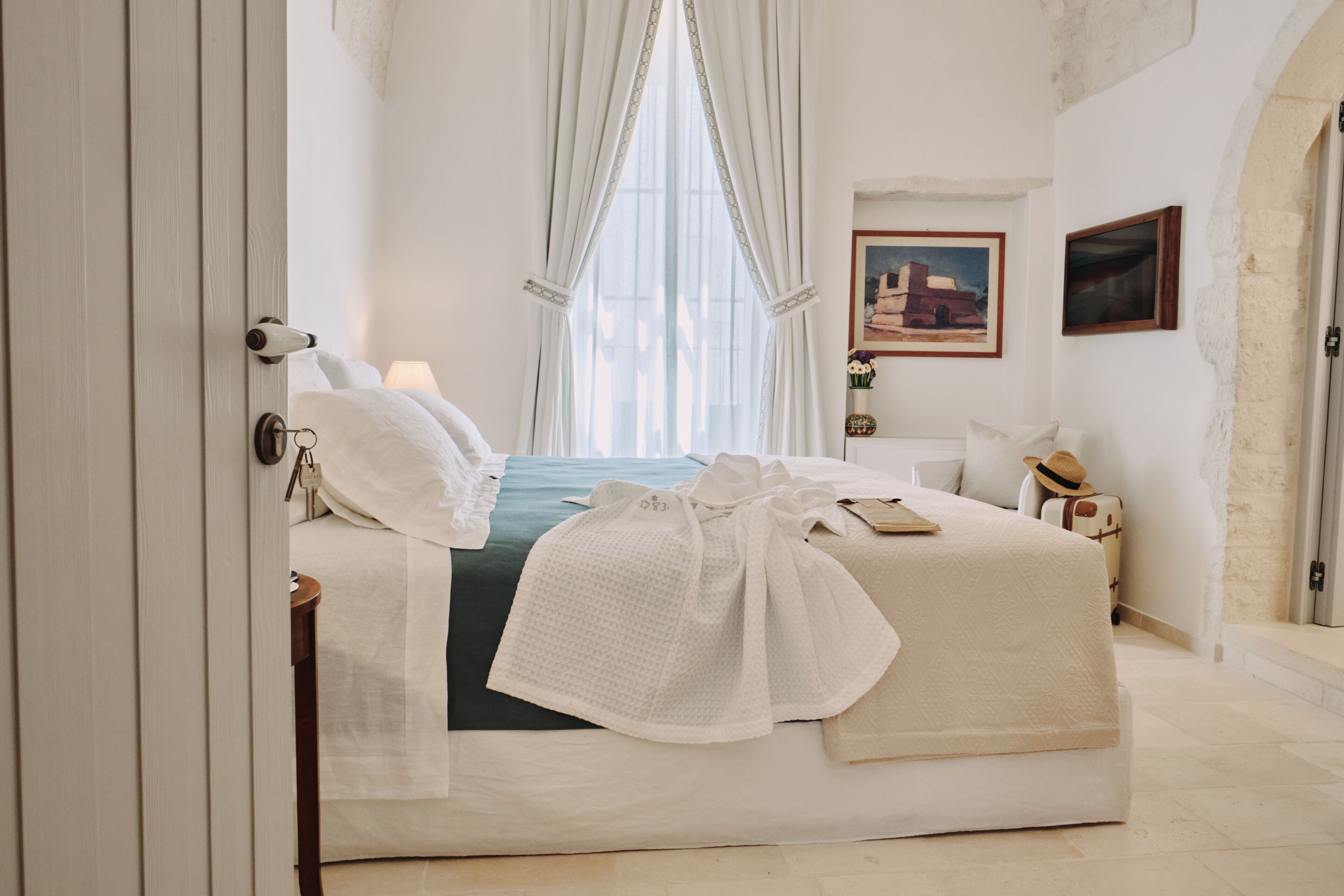 Una habitación de hotel con cama, mesita de noche, lámpara, ventana con cortinas y un cuadro en la pared.