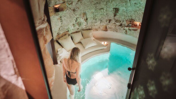 Een vrouw in een zwart zwempak staat aan de rand van een jacuzzi, met een stenen muur en een beige bank op de achtergrond.