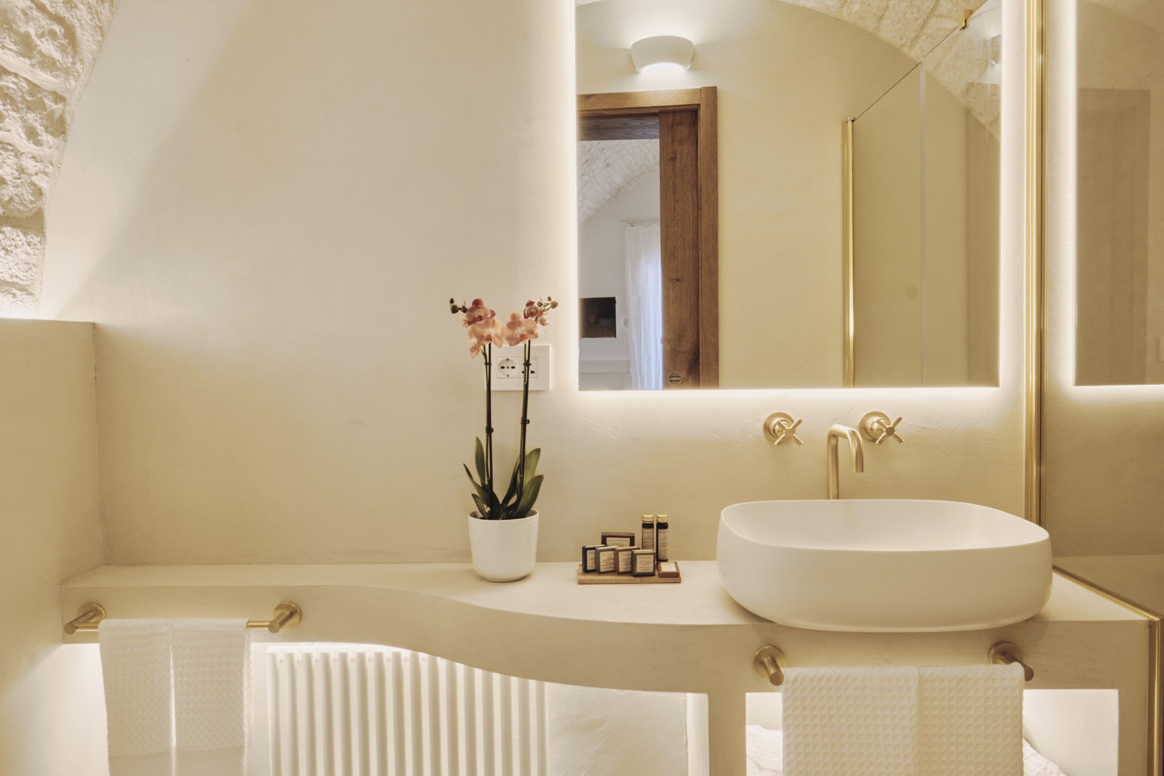 A modern bathroom with a large oval sink, two gold faucets, a white potted orchid, and a mirror above the sink.