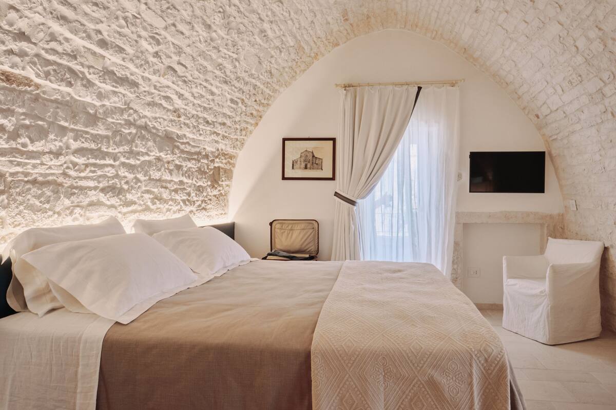 A bedroom with a stone wall, a bed with white bedding, a chair, and a flat-screen TV mounted on the wall.