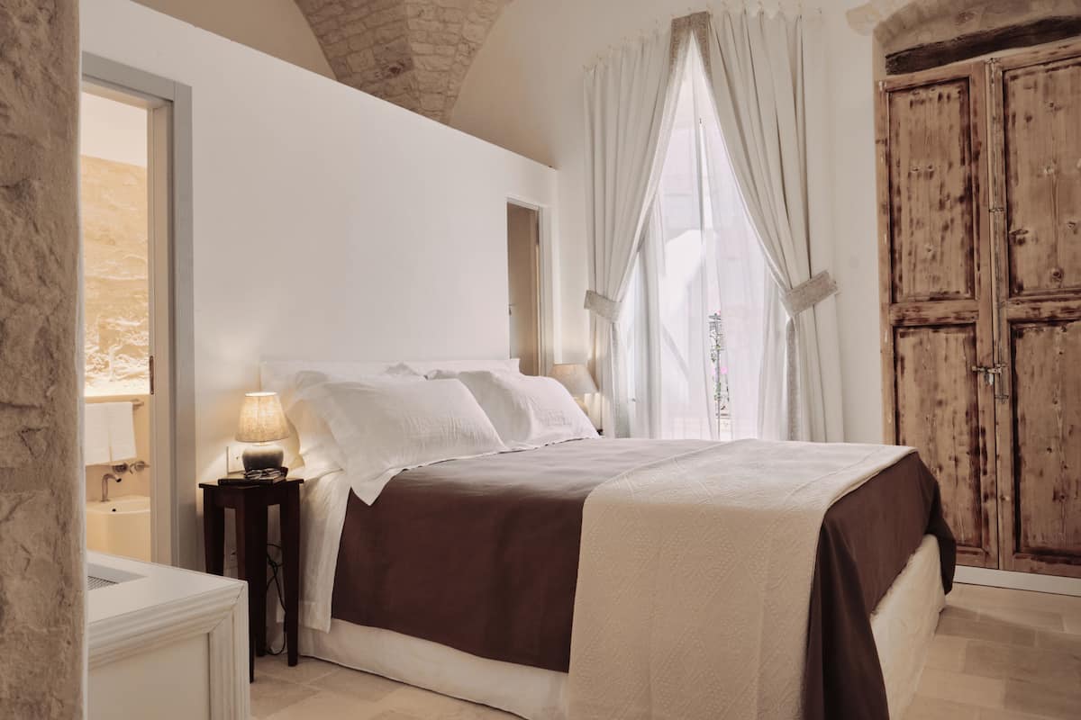 A bedroom with a large bed, a nightstand, a lamp, a wooden wardrobe, and a window with curtains.