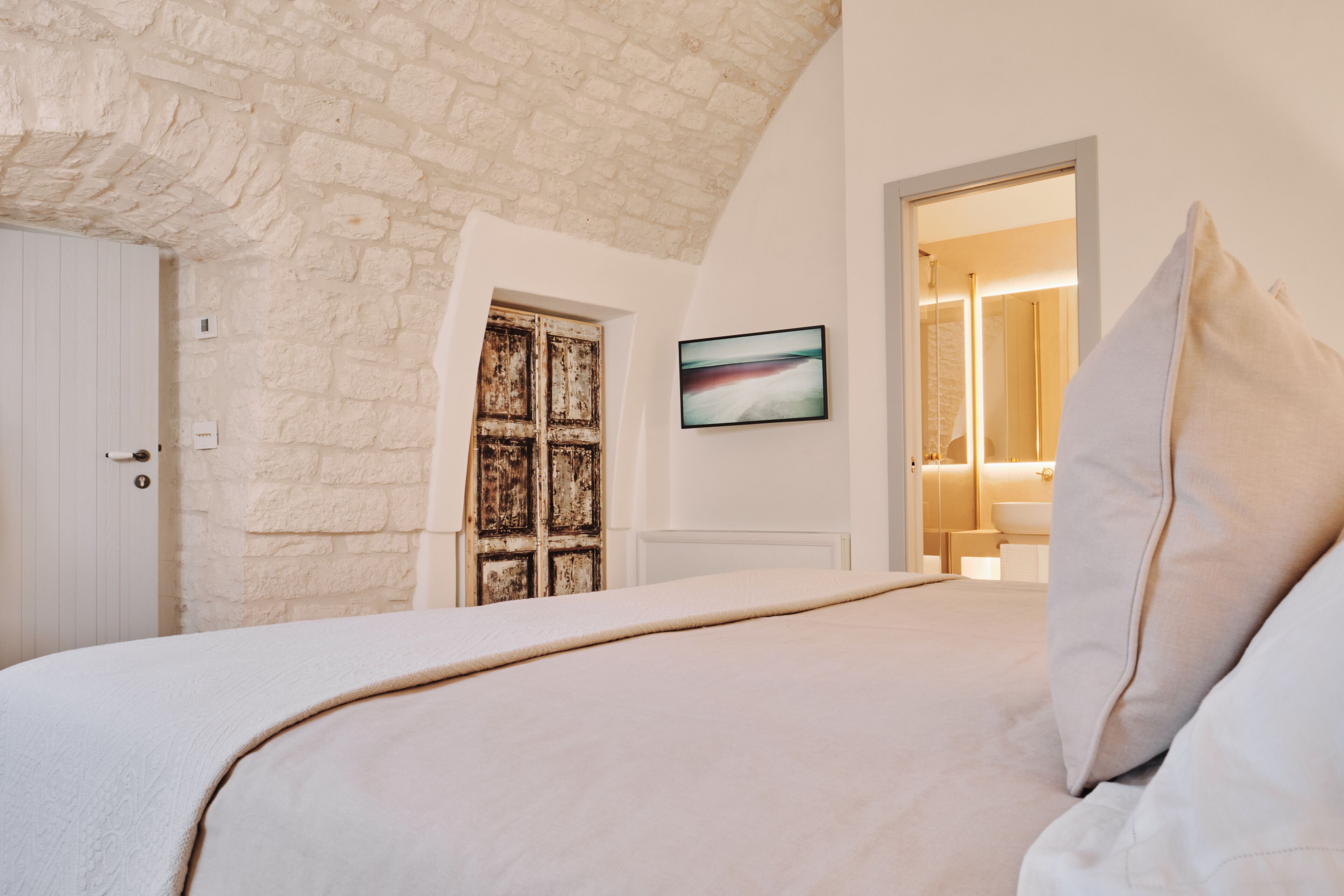 Une chambre avec un plafond en pierre, un lit recouvert de draps blancs, une porte en bois et un tableau encadré accroché au mur.