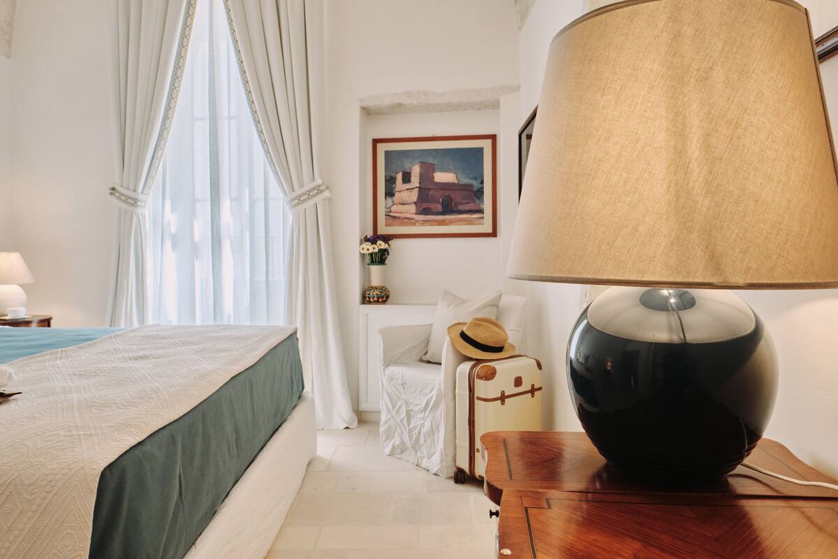 A hotel room with a bed, a lamp, a suitcase, and a painting on the wall.