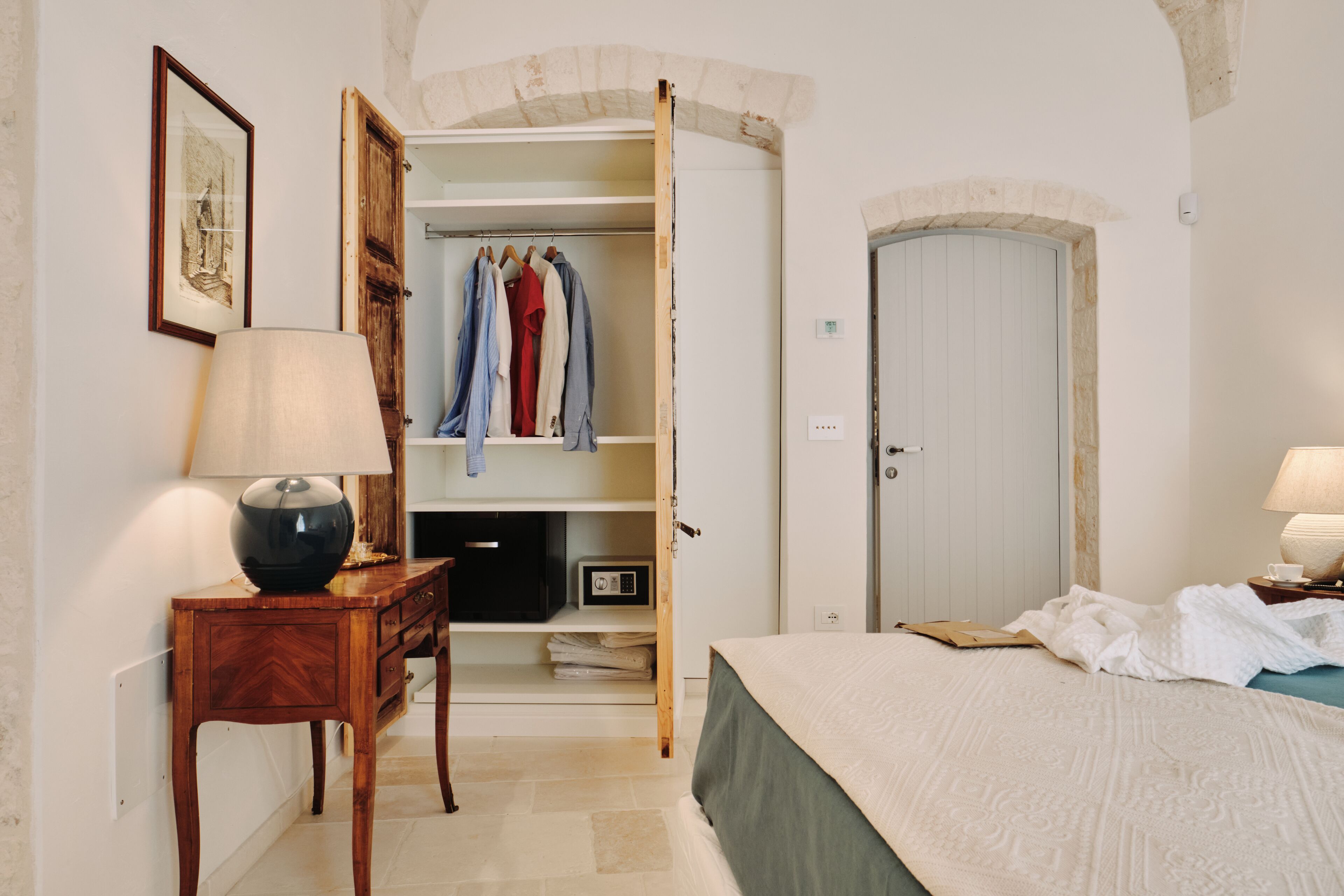 Une chambre à coucher avec un lit, une table de chevet avec une lampe, une commode en bois et une armoire où sont suspendus des vêtements.