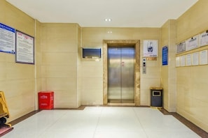 A hallway with a tiled floor, an elevator, a trash bin, and informational posters on the walls. - Youjia Hotel (Bijie)