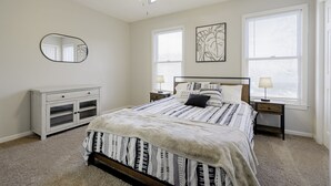 A bedroom with a bed, a ceiling fan, a nightstand with a lamp, a dresser, and a window with blinds. - Rosewood Townhouse (Columbia)