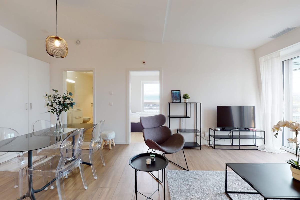 A modern living room with a glass dining table, clear chairs, a black coffee table, a grey sofa, a TV on a black stand, and a large window with curtains.