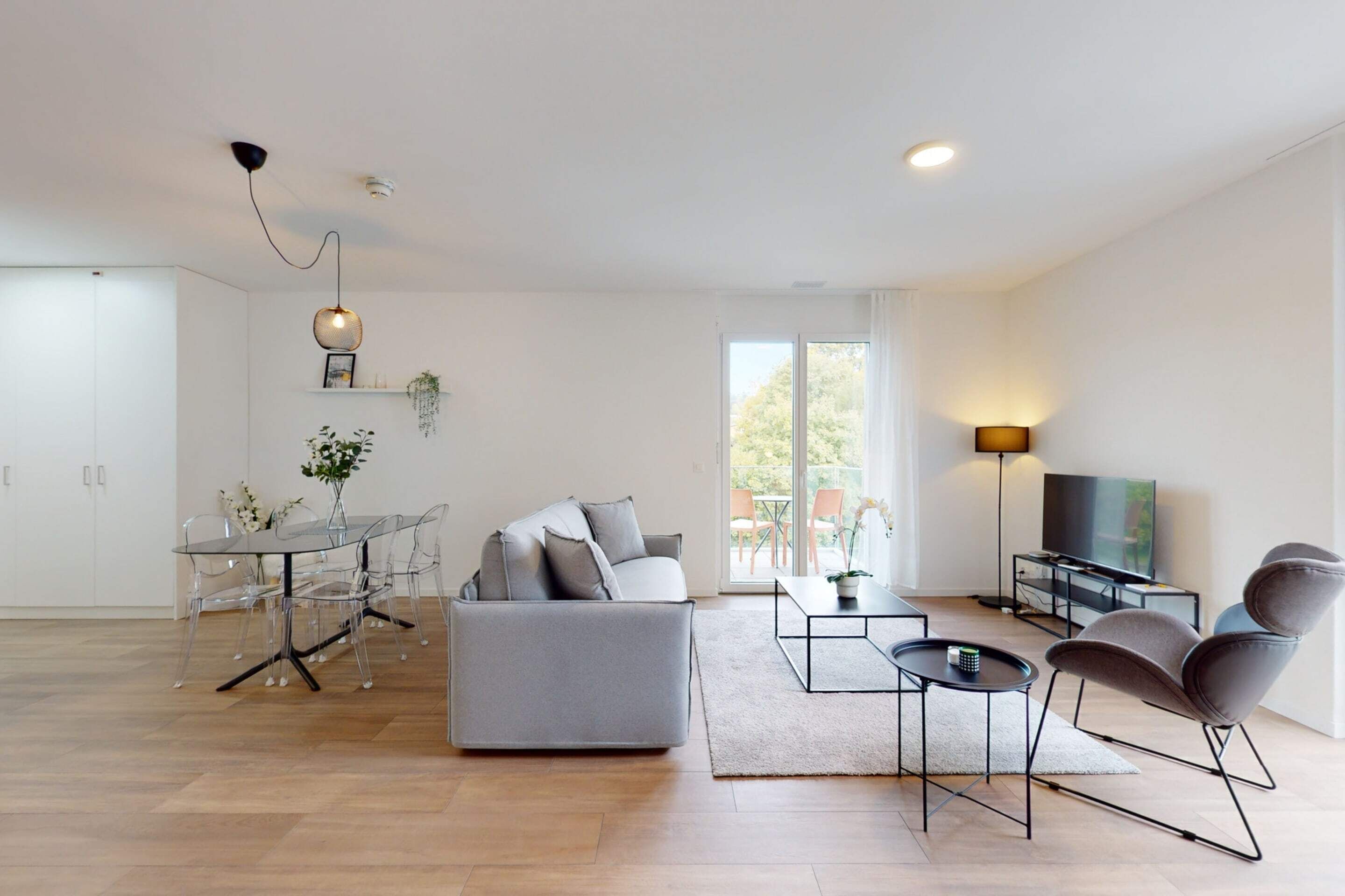 A modern living room with a grey sofa, a glass dining table, and a TV.