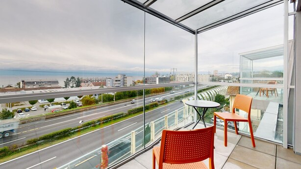 A balcony with a glass railing overlooking a busy road and distant buildings. - Lodges Hotel Morges (Morges)